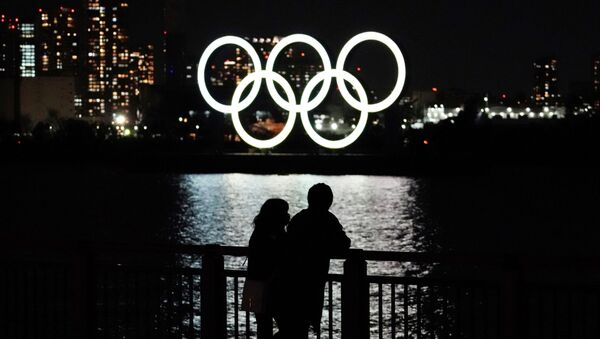 Люди в масках стоят на набережной на фоне гигантских светящихся олимпийских колец в Токио  - Sputnik Казахстан