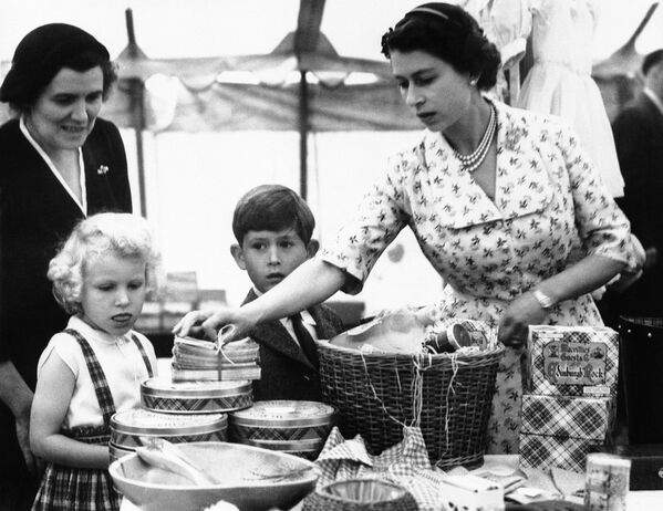 Королева Великобритании Елизавета II расставляет товары на распродаже в замке Абергелди, недалеко от замка Балморал в Шотландии, 20 августа 1955 года. Принцесса Анна внимательно наблюдает, пока принц Чарльз проявляет интерес к другим товарам. - Sputnik Казахстан