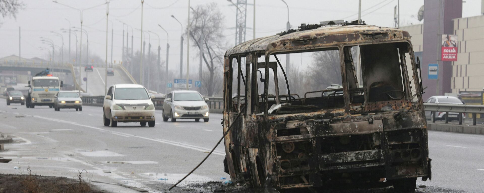 Машины двигаются по улице, на которой стоит разбитый и сожженный автобус - Sputnik Қазақстан, 1920, 29.01.2022