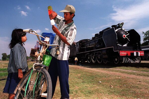 Камбоджиец готовит мороженое для маленькой девочки на фоне прибытия старого паровоза &quot;Пасифик 231&quot; на небольшой станции в 50 километрах к югу от Пномпеня, 6 февраля 2000 года.  - Sputnik Казахстан