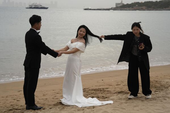 Женщина держит за волосы невесту, чтобы сделать свадебные яркие запоминающиеся фотографии на пляже в Циндао, Китай. - Sputnik Казахстан