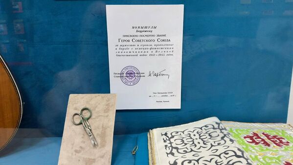 Батыр қолданған қайшы мен ол өз қолымен ойған оюлар. Осы заттардың жоғарысында орналасқан құжаттан Бауыржан Момышұлына Кеңес Одағының Батыры атағын беру туралы бұйрықтың мәтінін оқуға болады.  - Sputnik Қазақстан