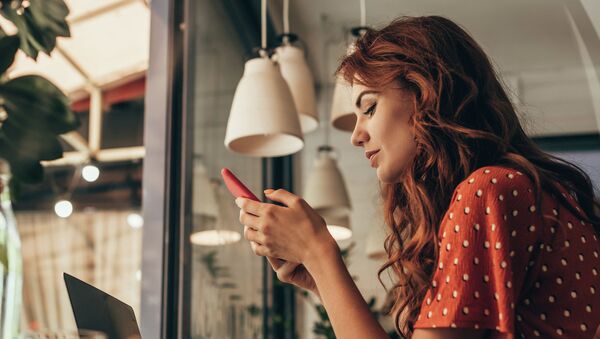 Рыжеволосая девушка в красном платье сидит в кафе и смотрит на свой смартфон - Sputnik Казахстан