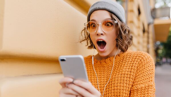 Сильно удивленная девушка в шапке и оранжевых очках смотрит на экран смартфона - Sputnik Казахстан